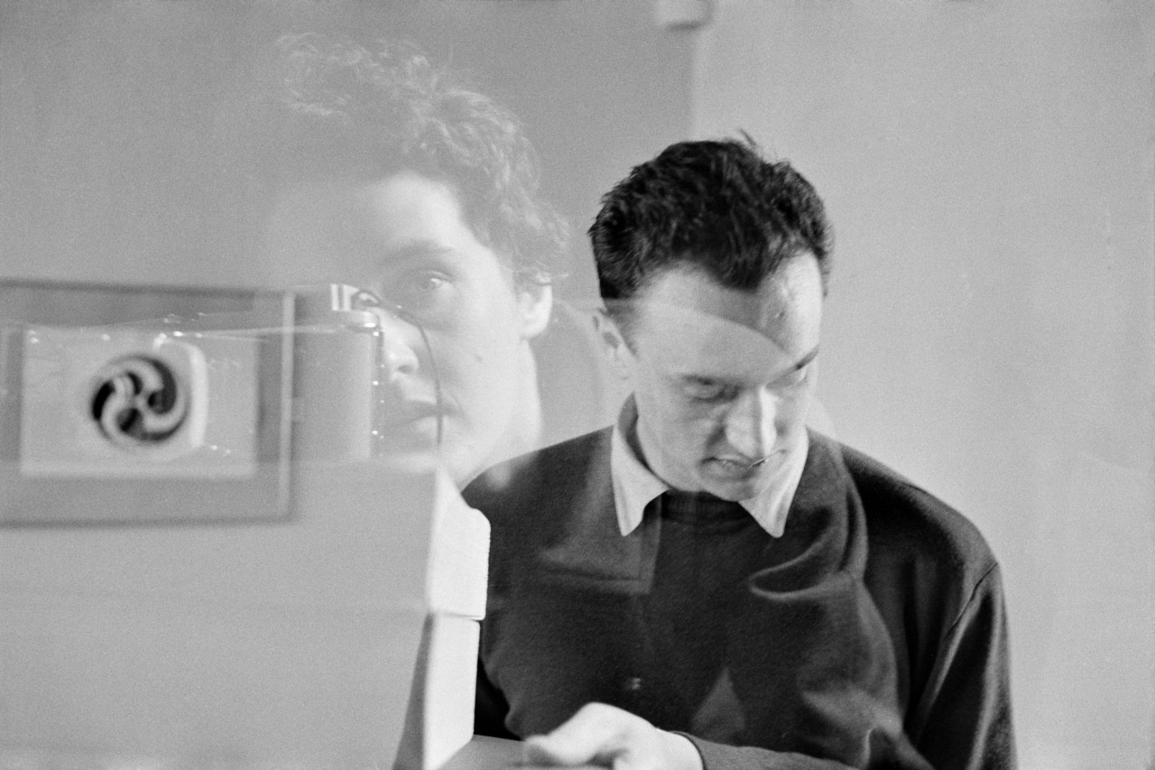 Rene Burri Robert Delpire, publisher, with double exposure of Inge Morath, Magnum photographer behind her Leica camera and Yin and Yang picture on wall. Paris, France. 1956. © Rene Burri | Magnum Photos